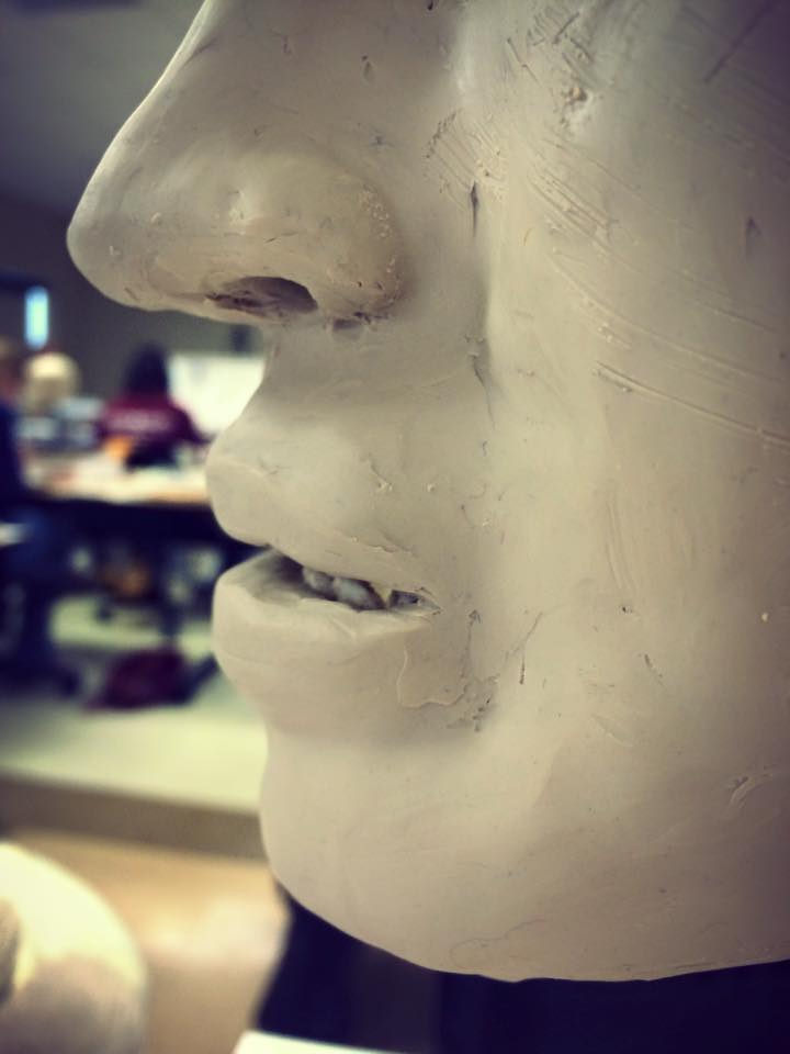 Close-up of sculpture showing nose and mouth. The sculpture's lips are open slightly to reveal teeth. There are hatch marks on the clay surface from using sandpaper to texturize it slightly. 