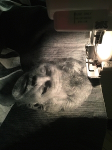 Fabric going under the foot of a serger machine. The fabric is printed with the black-and-white booking photo from my brother's police report. 